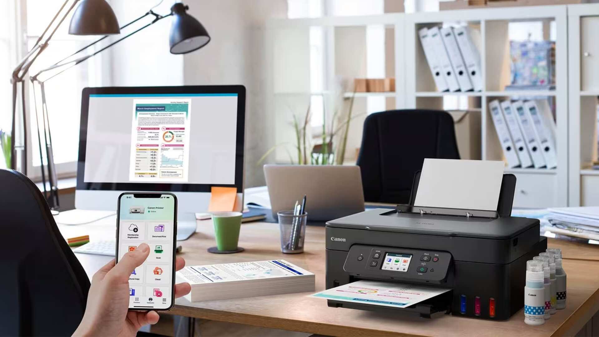 Black Canon printer on an office desk controlled by a smartphone app printing pages.