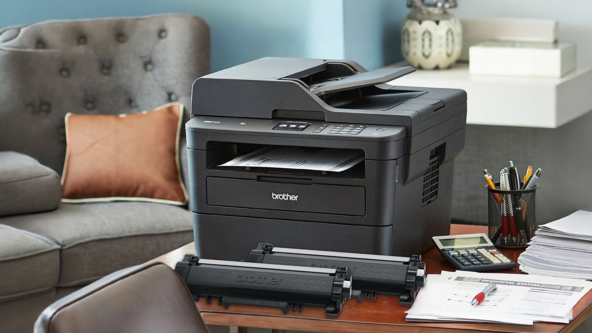 Black all-in-one laser printer with spare toner on a desk.