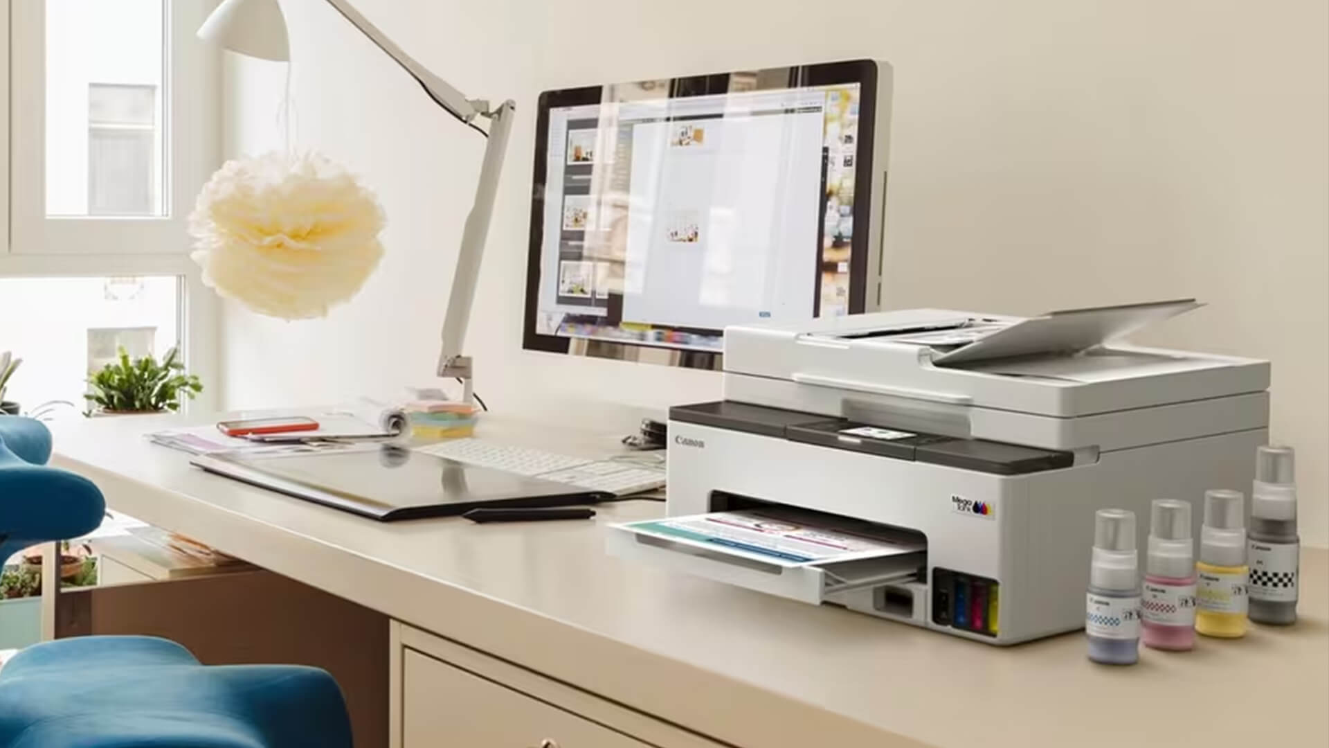 White Canon PIXMA MegaTank printer on a desk next to a computer monitor and ink bottles.