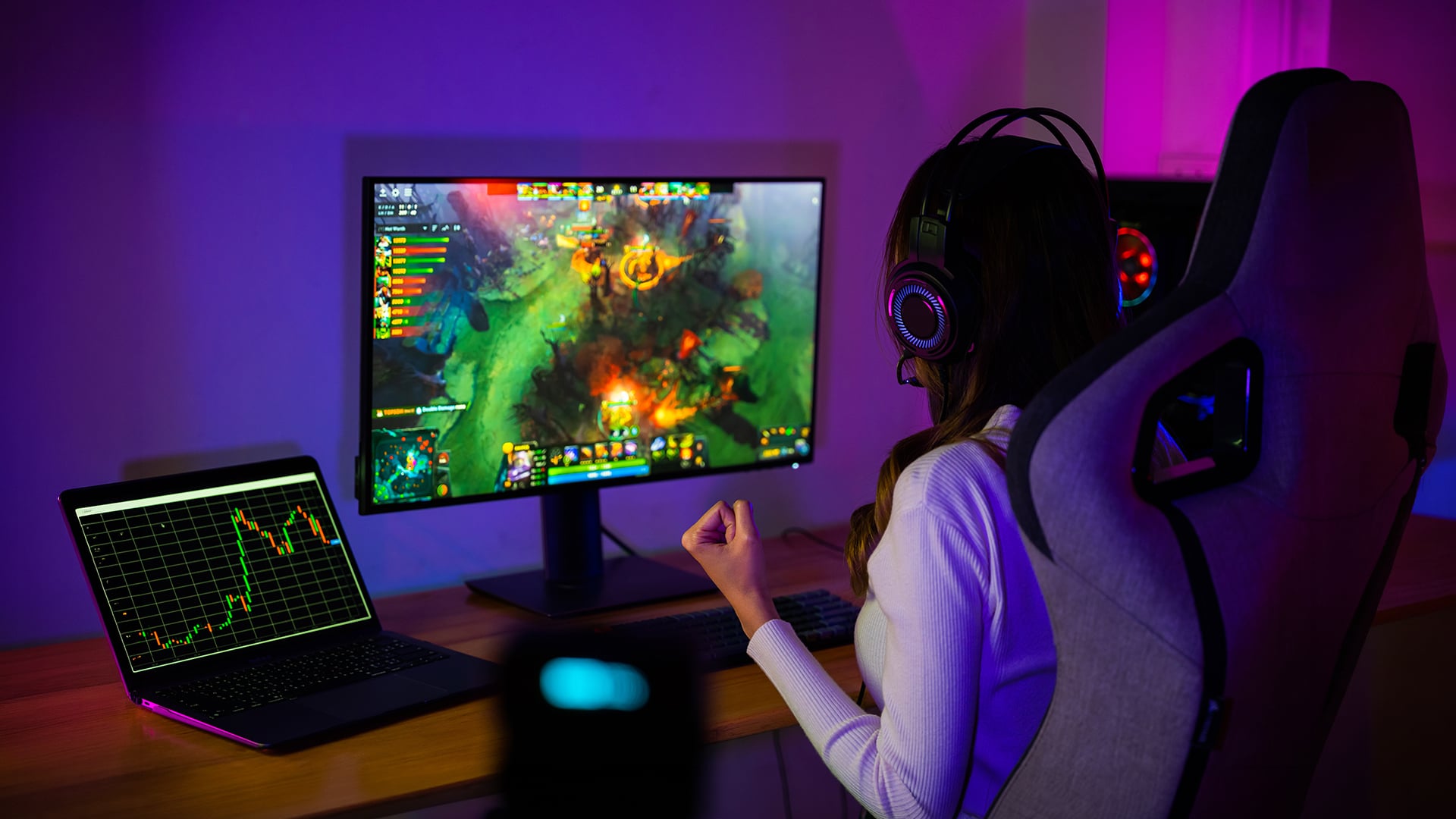 Female gamer at a desktop setup with a MOBA game on the monitor and financial charts on a laptop.