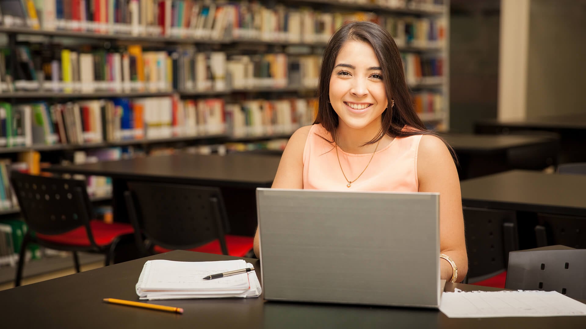 A student smiling at the camera while working on her laptop in a library.