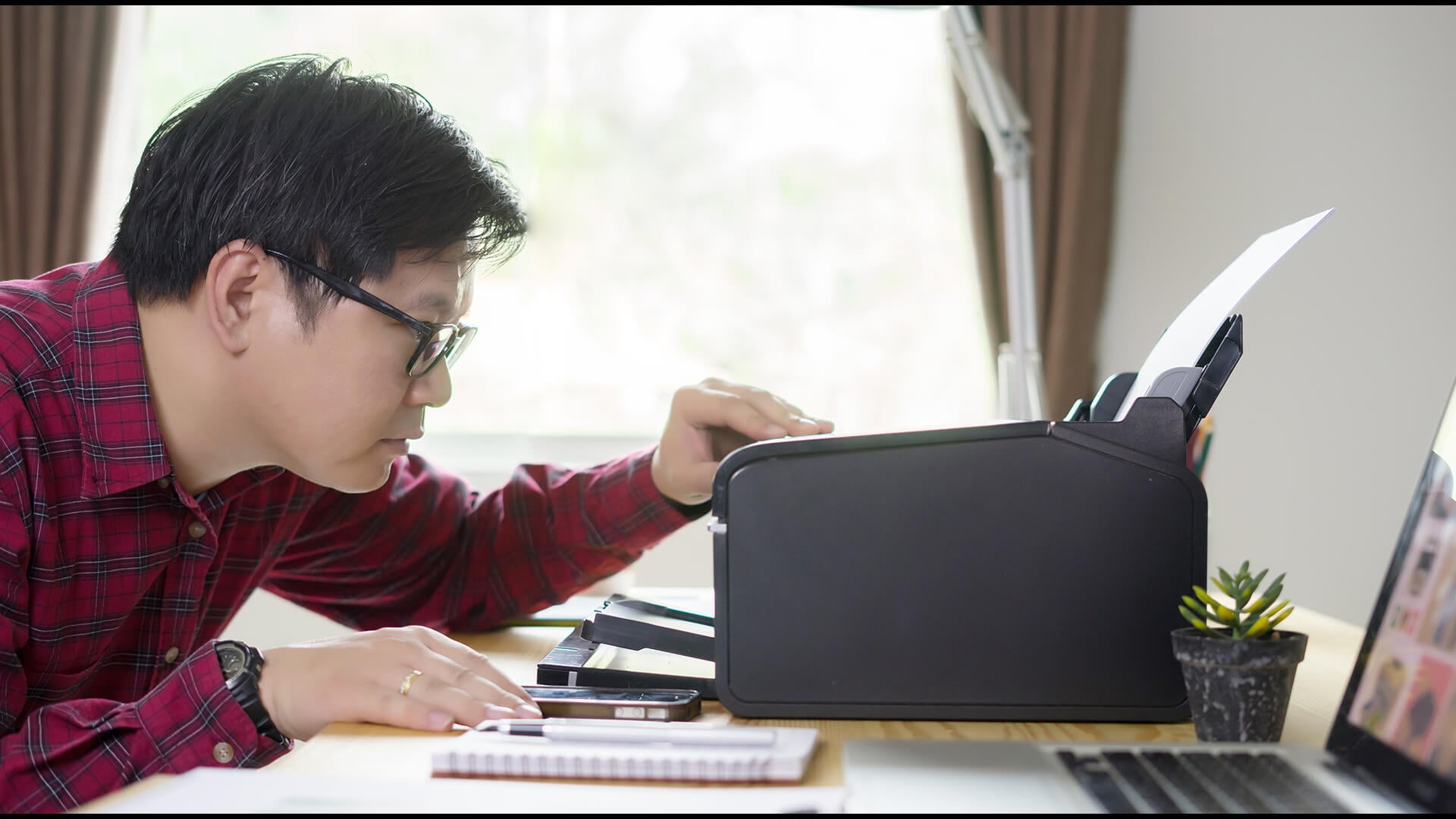 A man adjusting a black laser printer on a desk in a home office.