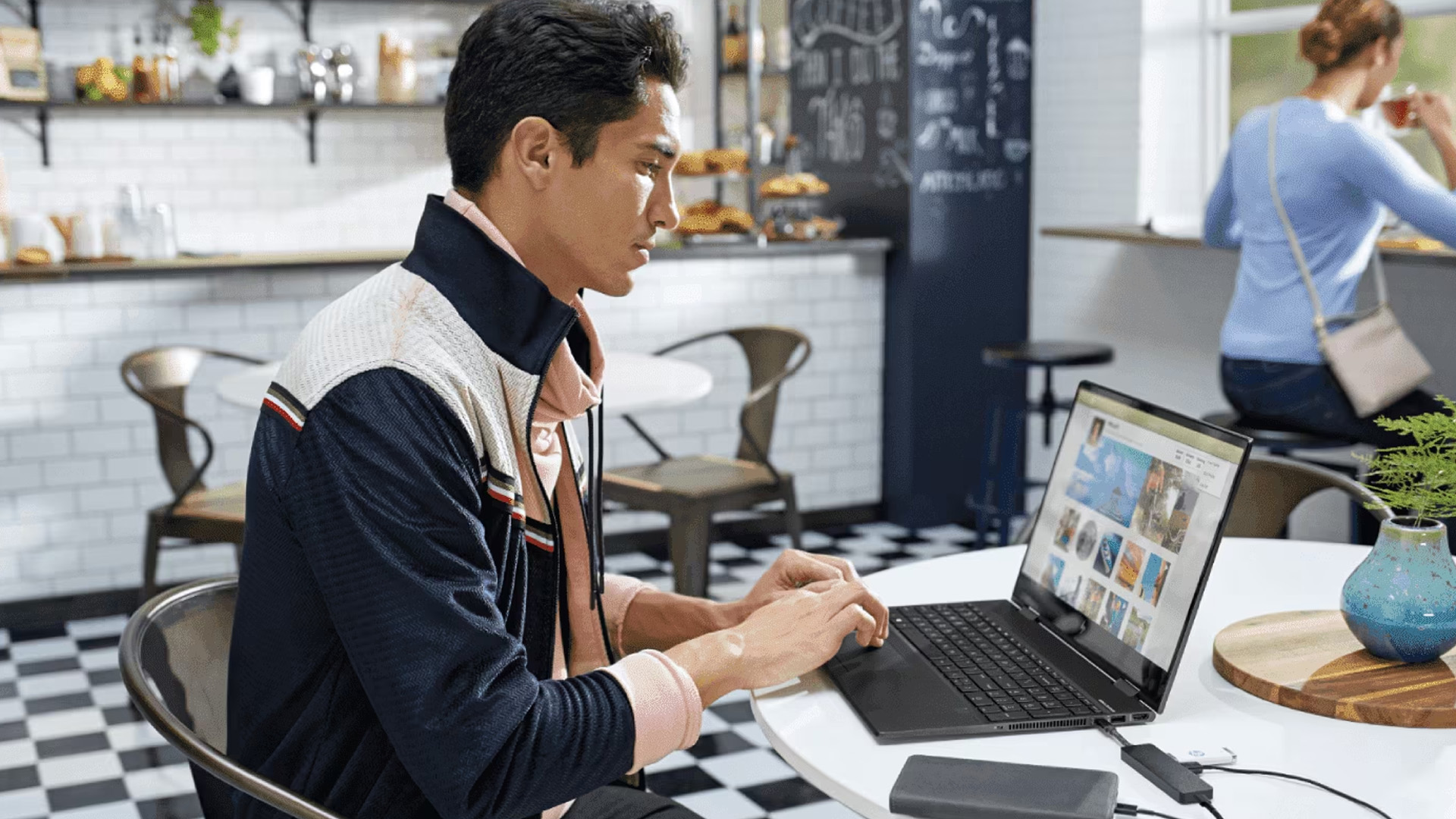Man typing on a plugged-in black HP laptop at a white cafe table.