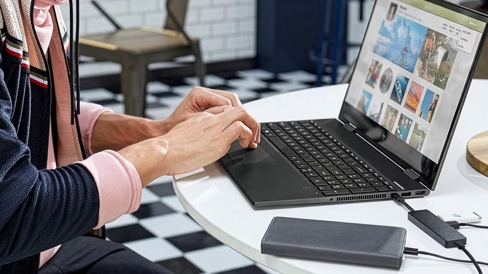 Person typing on a plugged-in black laptop sitting on a white table.