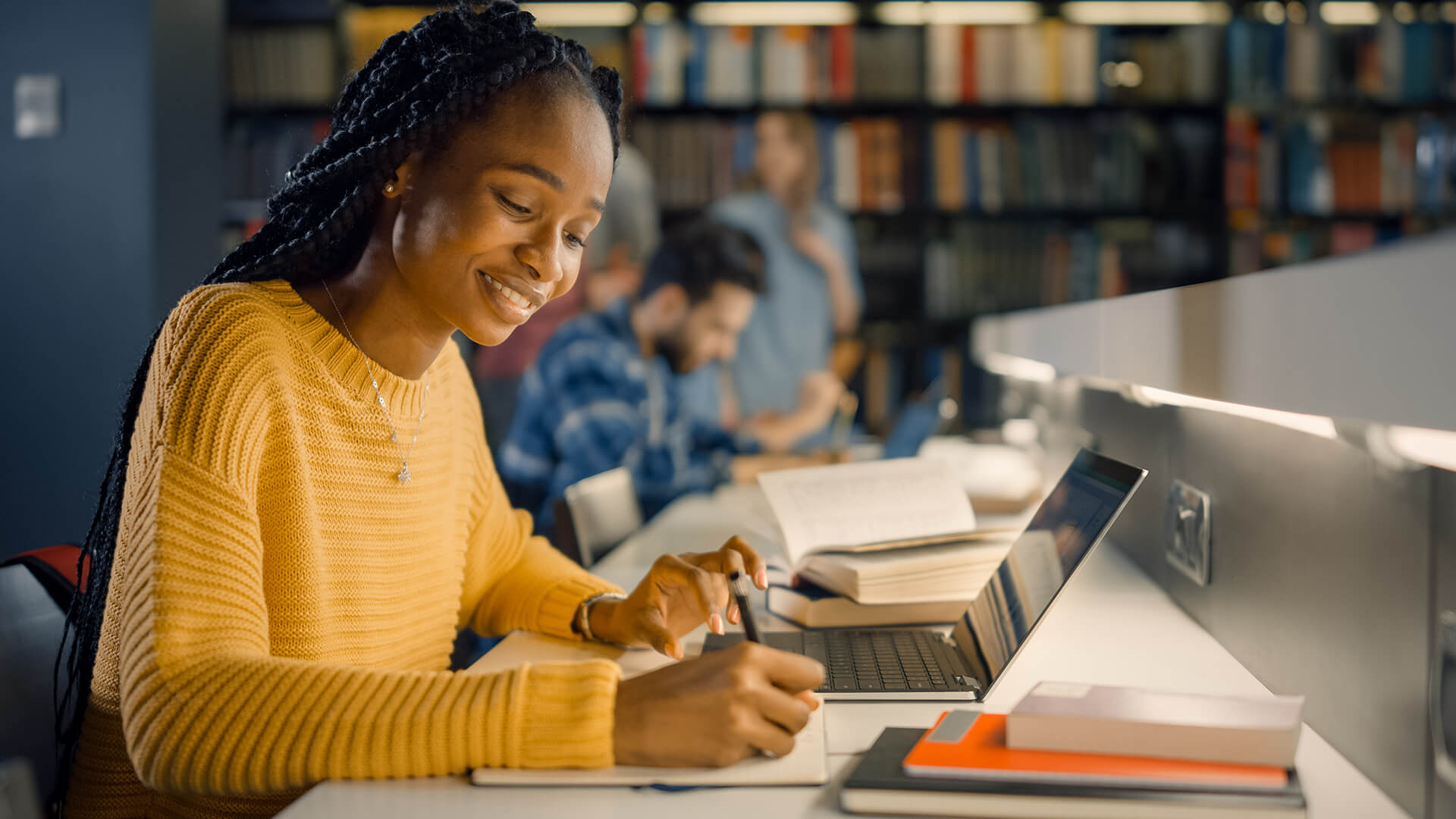 A female student taking notes next to her laptop inside a university library.
