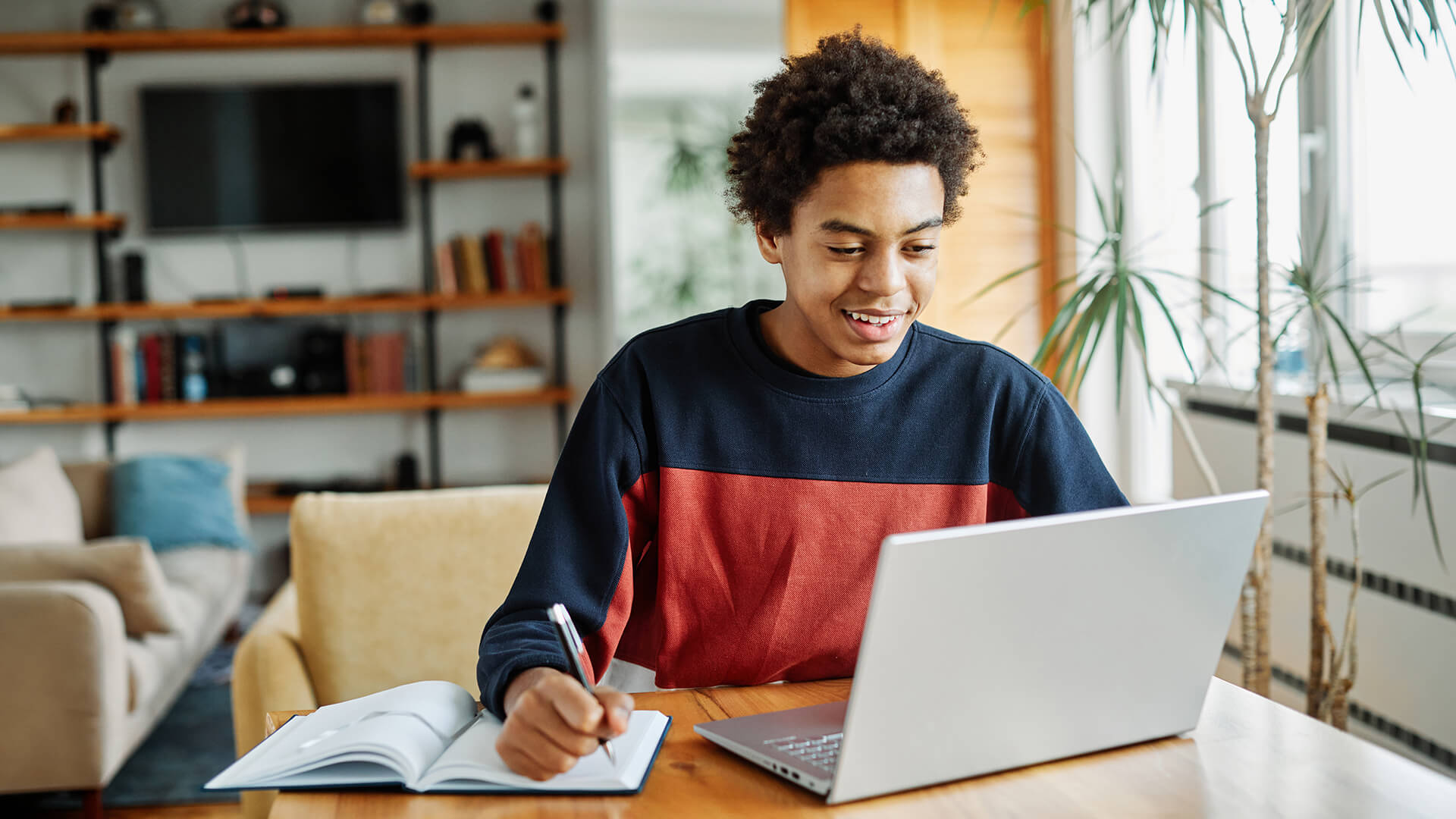 A young student smiling and writing in a notebook next to a laptop.