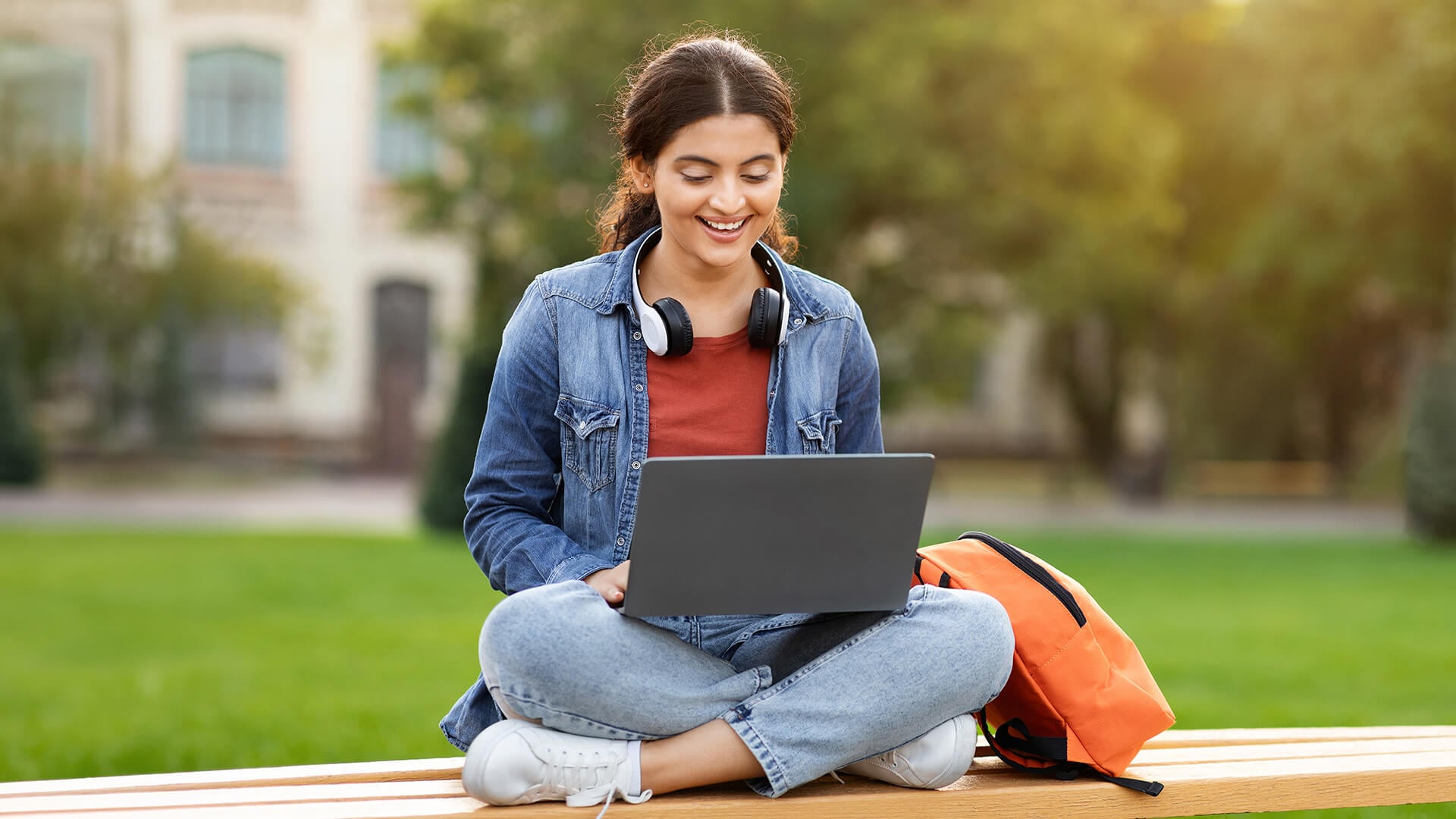 A smiling female student using her laptop while sitting on a campus bench.