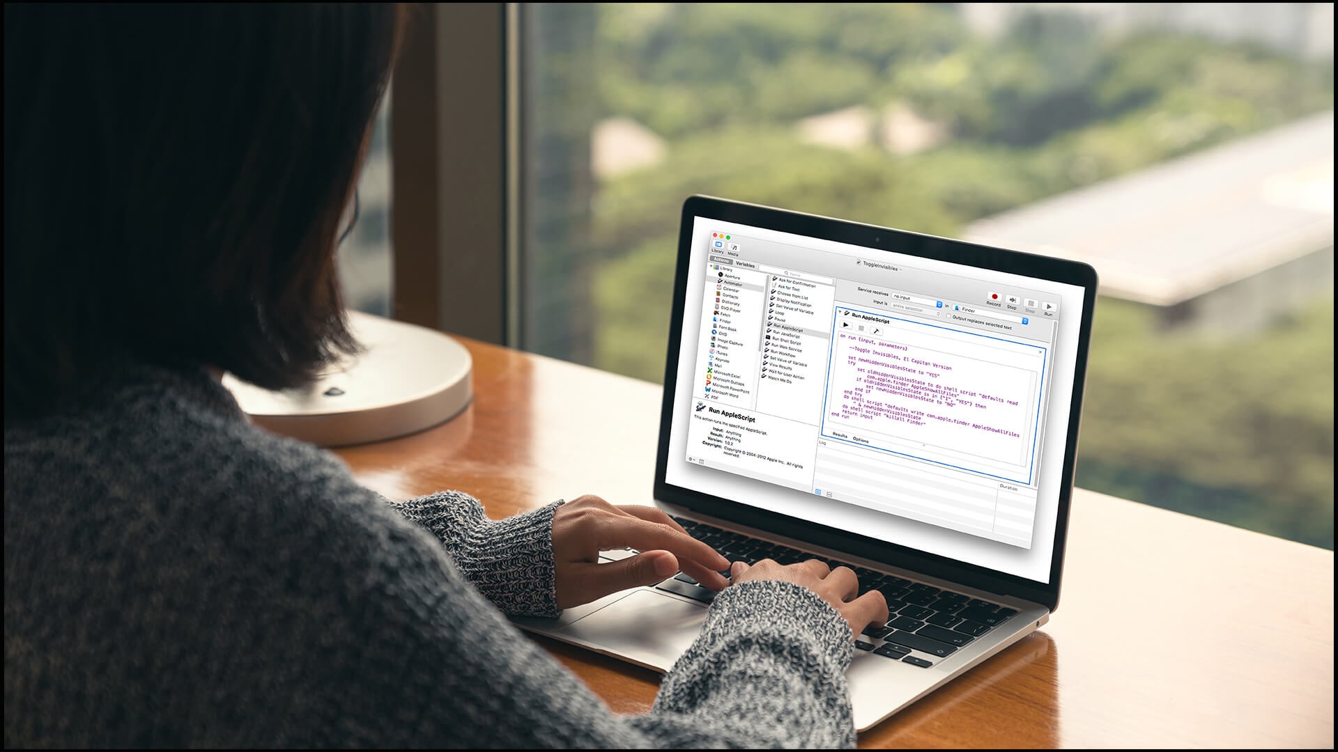 A woman typing code on a MacBook near a window in an office.