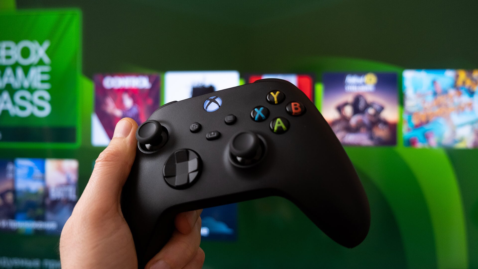 Hand holding a black Xbox controller with the Game Pass menu behind.